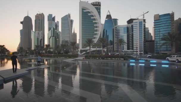 Zachodnia zatoka drapacze chmur w Doha Katar popołudniowe powiększenie zdjęć zrobione z parku Sheraton z fontanną na pierwszym planie, flagi Kataru, palmy i samochody poruszające się na ulicy Corniche - Materiał filmowy, wideo