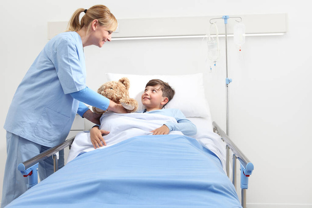 Lächelnde Krankenschwester gibt einem glücklichen Kind, das im Krankenhauszimmer im Bett liegt, einen Teddybär auf weißem Hintergrund mit Kopierplatz - Foto, Bild
