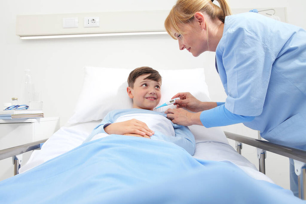 Happy child lying in bed in hospital room and nurse taking his temperature - Foto, afbeelding