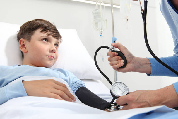 Dziecko leżące w łóżku w pokoju szpitalnym i pielęgniarka dokonująca pomiaru ciśnienia za pomocą sfigmomanometru i stetoskopu - Zdjęcie, obraz