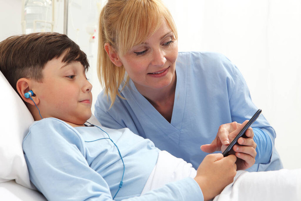 Happy child lying in bed in hospital room and smiling nurse using smartphone surfs the internet wearing earphones - Fotografie, Obrázek