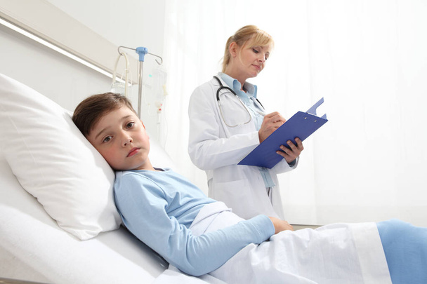 Sick child lying in bed in hospital room looking at camera with doctor beside him filling out his medical record with copy space - Фото, изображение