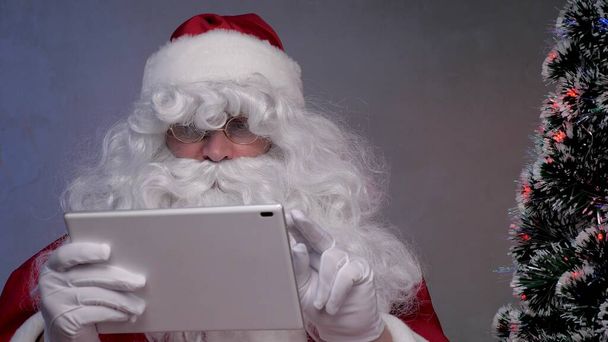 Joulupukki toimii etätyönä tabletin kanssa verkossa. Iloinen vanhempi mies joulupukin puku hymyillen ja katsoen moderni tabletti istuu sohvalla lähellä joulukuusi illalla huoneessa. Uusi vuosi Lomat - Valokuva, kuva