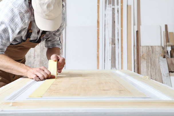Asztalos férfi, aki fával dolgozik ácsműhelyben, papírmaszkot ragasztva egy fából készült ajtóra, általános és sapkás. - Fotó, kép