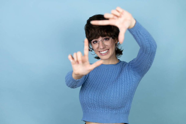 Young caucasian woman over isolated blue background smiling making frame with hands and fingers with happy face - Zdjęcie, obraz