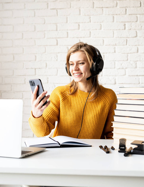 Distance learning. E-learning. Young smiling woman in black headphones studying online using laptop looking to mobile phone - 写真・画像