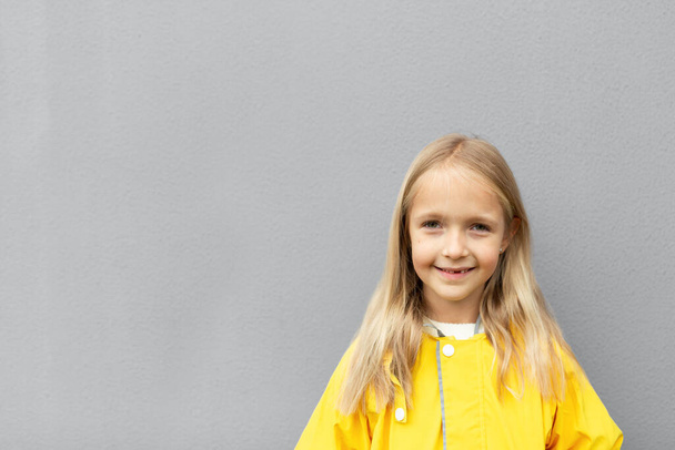Happy little caucasian girl 6-7 years old with blonde hair in yellow raincoat standing outdoor near gray wall. Colors of the year 2021 ultimate gray and illuminating - Photo, image
