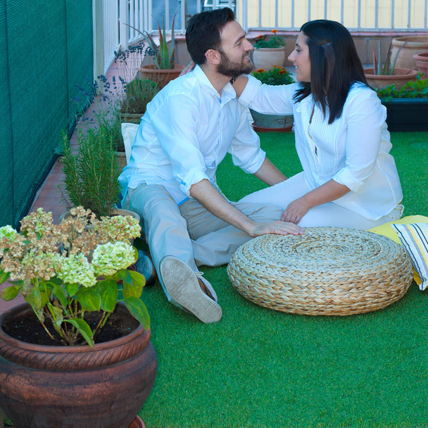 Молода пара, одягнена в біле вбрання, готує терасу до того, щоб вона насолоджувалася домашнім днем валентинки, використовуючи рослини, траву та подушки. - Фото, зображення