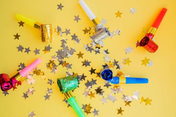 Flat lay of whistles and stars confetti.Birthday celebration attributes. Party horns, shiny confetti and other items on bright yellow background. party accessories - Photo, Image