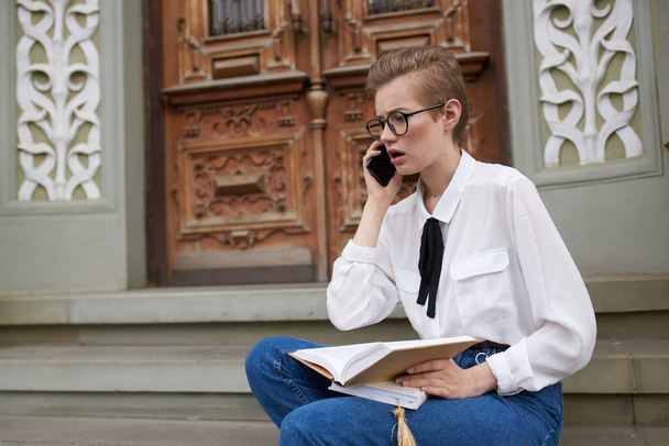 female student sits on the steps near the building with a book in her hands and a mobile phone - Foto, Imagen