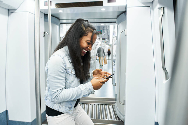 Портрет молодой женщины в маске, использующей свой мобильный телефон в вагоне метро. Улыбающаяся афроамериканка со смартфоном. - Фото, изображение