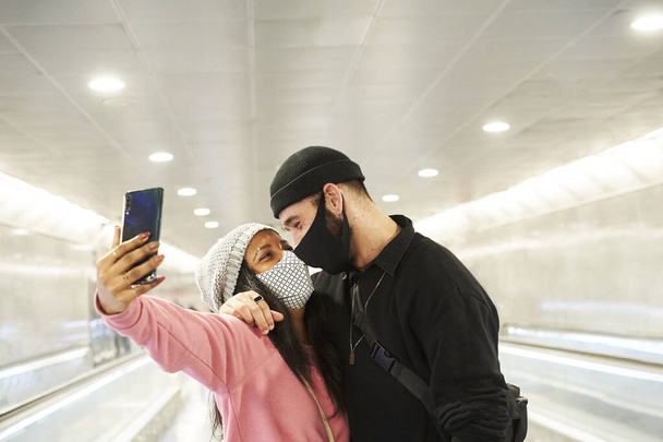 Młoda międzyrasowa para kochanków w maskach i wełnianych kapeluszach robiących selfie w metrze lub korytarzu lotniska. - Zdjęcie, obraz