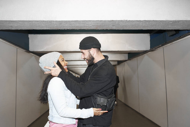 Joven pareja interracial de amantes con máscaras faciales en un corredor subterráneo de metro en los brazos del otro por un antiguo corredor de transporte público mirándose y disfrutando de su amor. - Foto, Imagen