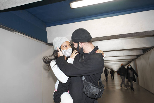 Joven pareja interracial de amantes en máscaras faciales y sombreros de lana se aman entre sí en la multitud en un corredor subterráneo del metro en los brazos del otro por un antiguo corredor de transporte público. - Foto, Imagen