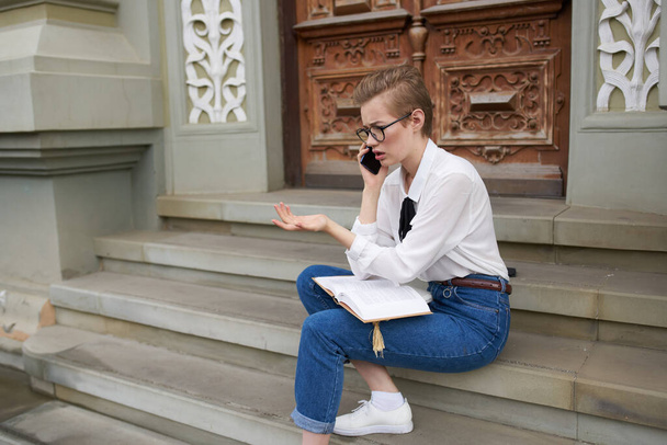 mujer en jeans camisa y zapatillas de deporte se sienta en las escaleras cerca del edificio del instituto de educación teléfono - Foto, Imagen