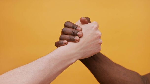 Schwarze und weiße Hände, die sich isoliert auf dem orangefarbenen Hintergrund zusammenhalten. Nahaufnahme - Foto, Bild