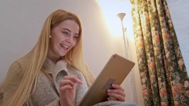 Счастливая улыбающаяся молодая женщина сидит на диване, друг на видео звонит с помощью планшета - Кадры, видео