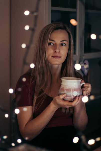gondos pillantás ki az ablakon egy ünnepélyes pillanatban. Érzelmi egyensúlyhiány. Egy fiatal nő egy csésze tea mellett töpreng a sorsán. Az európai nemzetiségű lány ül a karácsonyi ünnepi asztalnál. - Fotó, kép