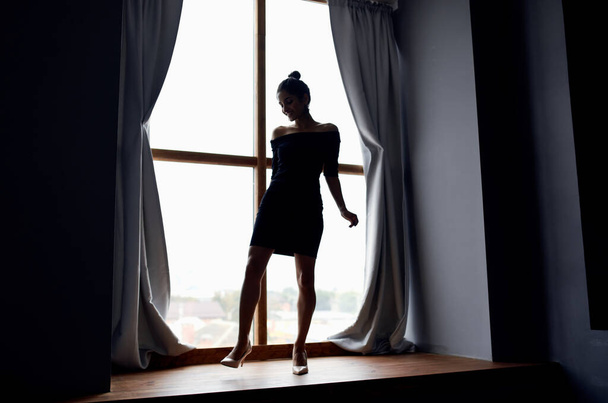 背景のカーテンの近くの窓辺の短いドレスを着た女性のシルエット - 写真・画像