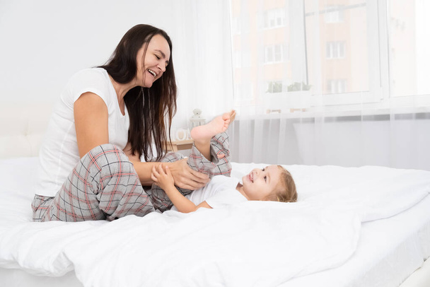 взрослая мать играет с маленькой активной дочерью в постели дома, весело, активность с детьми. - Фото, изображение