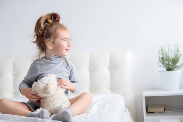 klein meisje in grijze coltrui met teddybeer zittend op bed glimlachend. - Foto, afbeelding