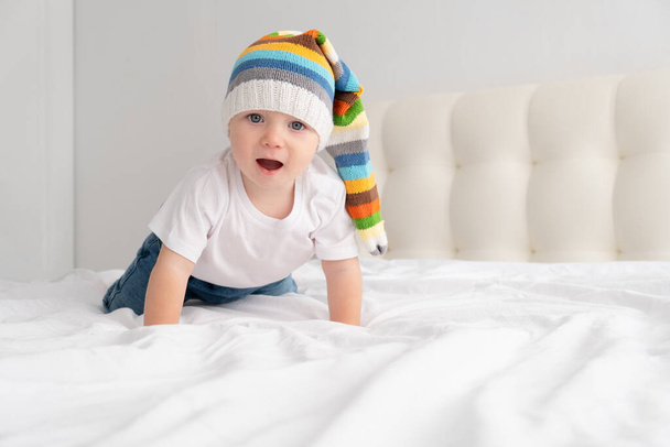 grappig baby jongen in grappige kleurrijke hoed glimlachen en liggen op een wit beddengoed thuis.  - Foto, afbeelding