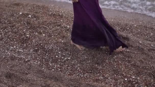 a piedi nudi donna in un lungo abito viola passeggiate lungo la spiaggia di ghiaia - Filmati, video