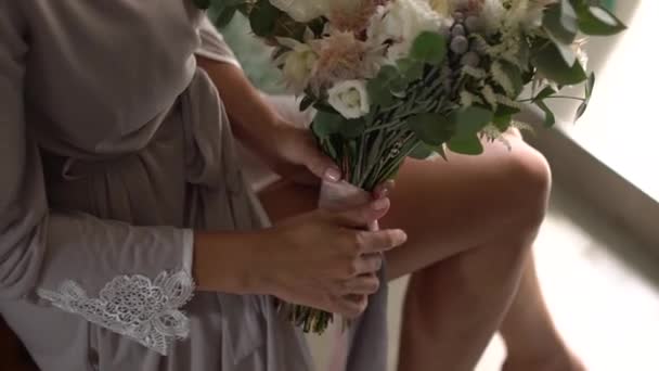 A woman in a silk robe holds a bridal bouquet on her lap and plays with long ribbons - Footage, Video