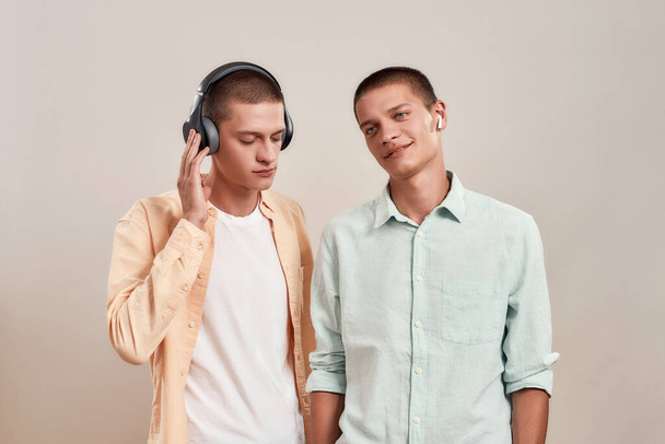 Two young caucasian twin brothers in casual wear listening music in headphones and wireless earphones, while standing isolated on beige background - Photo, Image