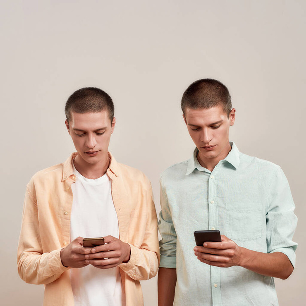 Two young caucasian twin brothers in casual wear using smartphones while standing isolated over beige background - Foto, imagen