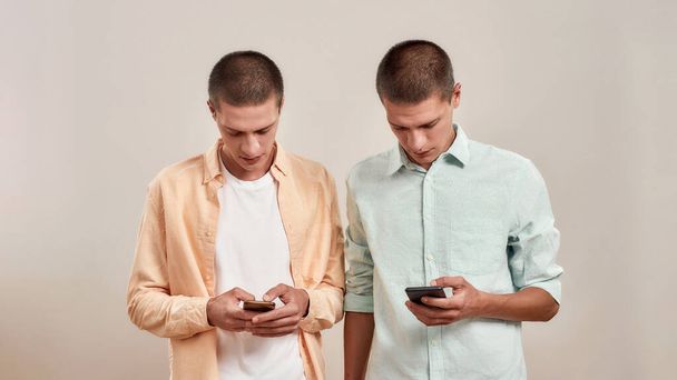 Two young caucasian twin brothers in casual wear using smartphones while standing isolated over beige background - Valokuva, kuva