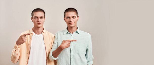 Two young twin brothers in casual wear pointing to the right and looking at camera while standing together isolated over beige background - Photo, image