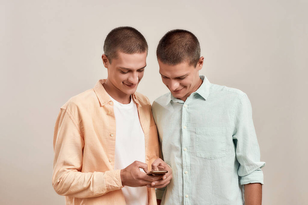 Two cheerful young twin brothers in casual wear using smartphone app while standing together isolated over beige background - Фото, изображение