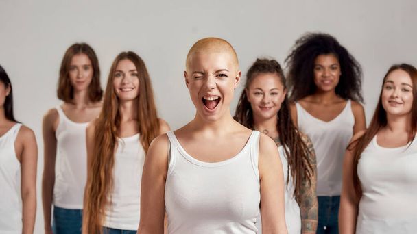 Portrait of cheerful young caucasian woman with shaved head in white shirt winking at camera. Group of diverse women standing isolated over grey background - 写真・画像