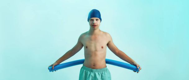 Disabled boy with Down syndrome in swimming cap looking at camera, holding foam noodle while posing isolated over turquoise background. Swimming rehabilitation concept - Photo, image
