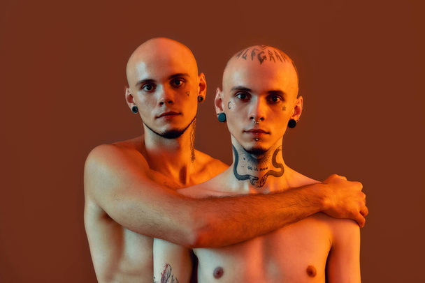 Portrait of young half naked twin brothers with tattoos and piercings looking at camera, posing together, standing isolated over orange background - Фото, зображення