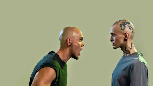 Portrait of two young men, twin brothers with tattoos and piercings arguing, shouting, standing face to face isolated over light green background - Photo, image