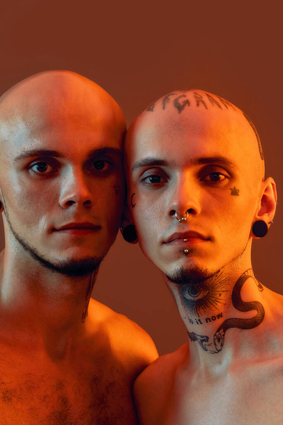 Close up portrait of young half naked twin brothers with tattoos and piercings looking at camera, posing together, standing isolated over orange background - Фото, зображення