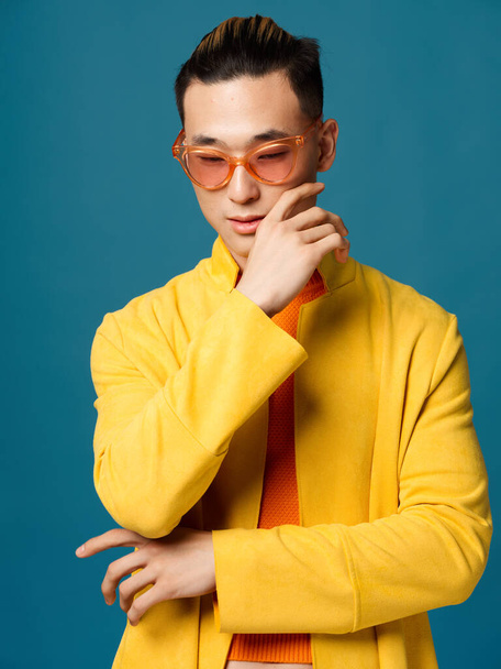 Hombre asiático con chaqueta amarilla sobre fondo azul y gafas en la cara, peinado - Foto, imagen