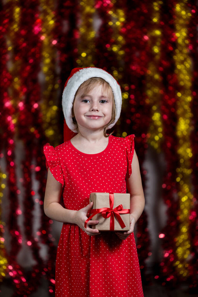 Очаровательная маленькая девочка в Санта-Шляпе держит новогодний подарок с мечтательным лицом, смотрящим в камеру. - Фото, изображение
