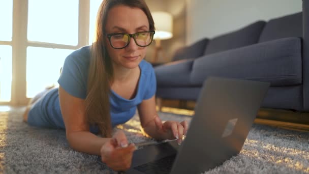 眼鏡をかけた女性が床に横になっていて、クレジットカードやノートパソコンを使ってオンラインで購入しています。オンラインショッピング、ライフスタイル技術 - 映像、動画