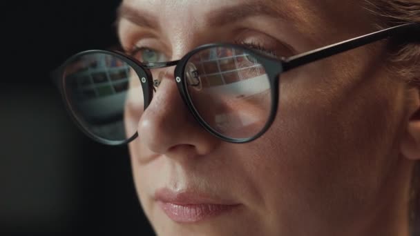 Woman in glasses looking on the monitor and surfing Internet. The monitor screen is reflected in the glasses. Work at night. Home Office. Remote work - Footage, Video