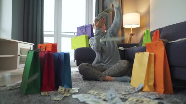 Szczęśliwa kobieta siedzi na dywanie w przytulnym pokoju wśród toreb na zakupy i zarabia pieniądze na deszczu z amerykańskich banknotów dolarowych - Materiał filmowy, wideo