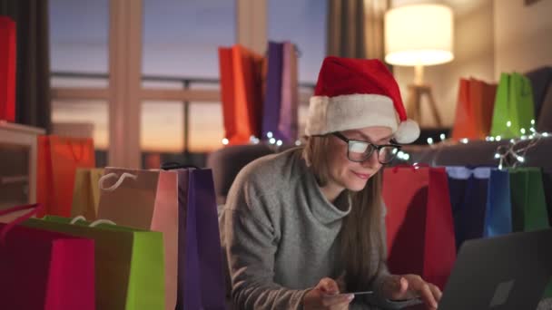 Mujer feliz con gafas con un sombrero de Santa Claus está acostado en la alfombra y hace una compra en línea con una tarjeta de crédito y un ordenador portátil. Bolsas de compras alrededor. - Metraje, vídeo