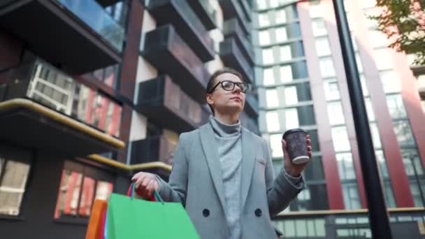 Formell gekleidete Frau spaziert nach dem Mittagessen mit Kaffee zum Mitnehmen und Einkaufstüten durch das Geschäftsviertel - Filmmaterial, Video