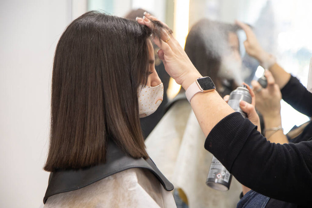 Hairdresser combing a brunette teenager with a lacquer mask after cutting her hair. In the mirror you can see the reflection of both of them. - Фото, изображение