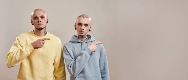 Two young twin brothers in casual wear with tattoos and piercings pointing to the right, looking at camera while standing together isolated over beige background - Foto, Imagem