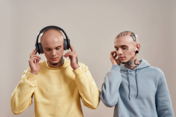 Creative people. Two young twin brothers in casual wear with tattoos and piercings listening music in headphones and wireless earphones - 写真・画像