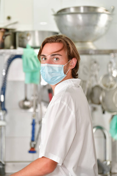 Close up of a young male chef using a surgical mask looking at the camera on an out of focus kitchen background. Cooking and safety concept. - Foto, Imagem