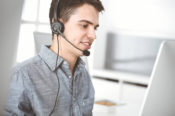 Casual dressed young man using headset and computer while talking with customers online. Call center, business concept - Foto, Imagen